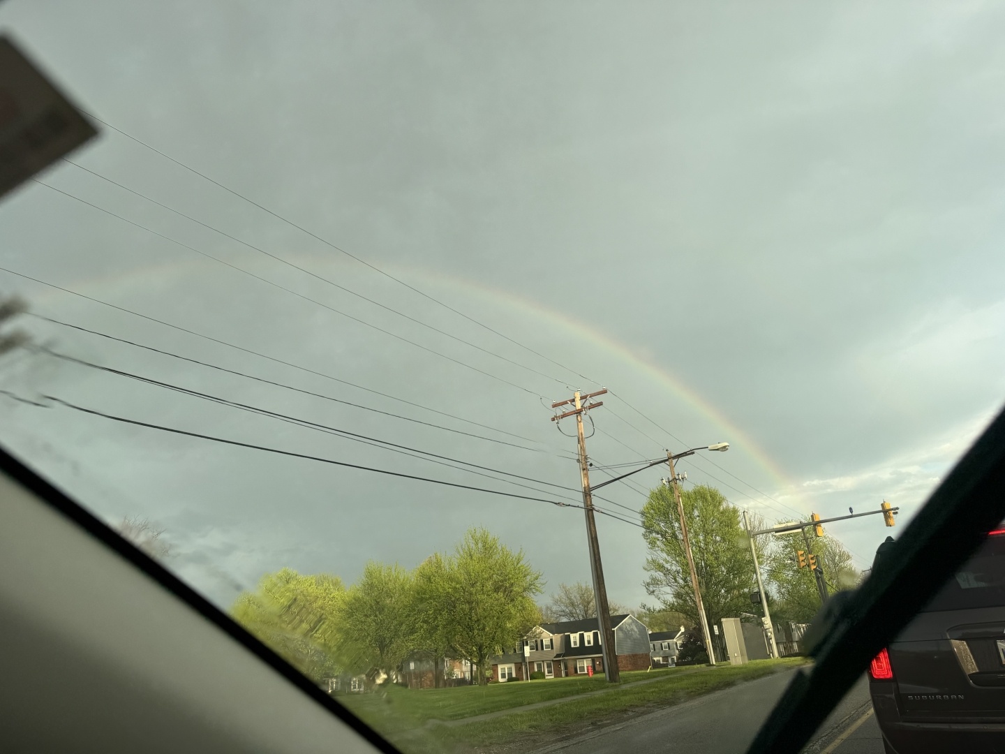 An arched rainbow appears against a backdrop of gray clouds, with green trees and residential houses visible below. Utility poles and power lines are also present in the scene, viewed from inside a vehicle.