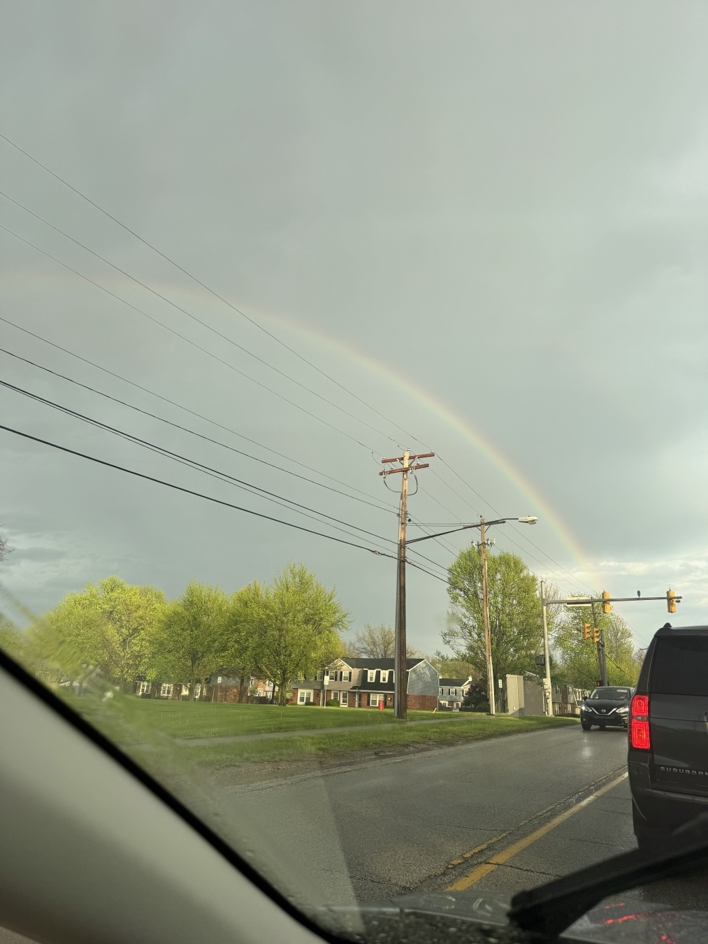 A rainbow arcs over a suburban street scene, with green trees and houses in the background. Power lines run across the sky, and vehicles are stopped at a traffic light. The sky appears cloudy, suggesting recent rain.