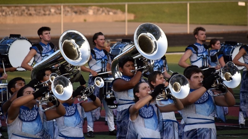 A drum corps performs on a field, featuring musicians in blue and white uniforms playing brass instruments. Drummers are positioned in the background. The scene captures the energy and coordination of the group.