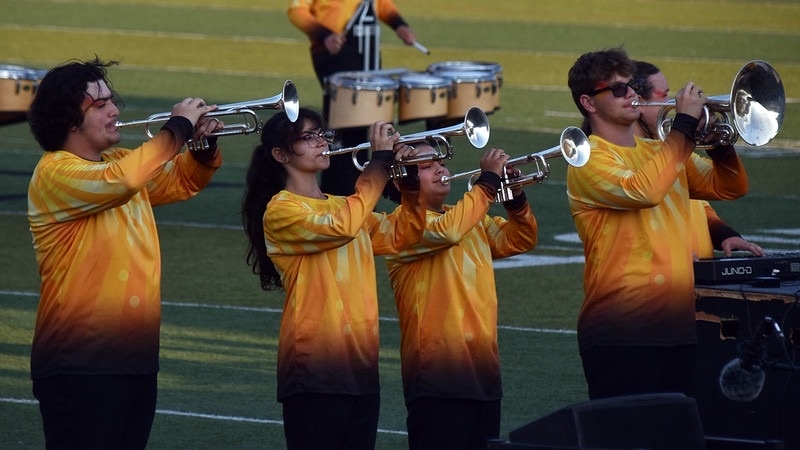 A group of four musicians in matching yellow and orange gradient shirts play trumpets on a field. In the background, there are drums and other performers.