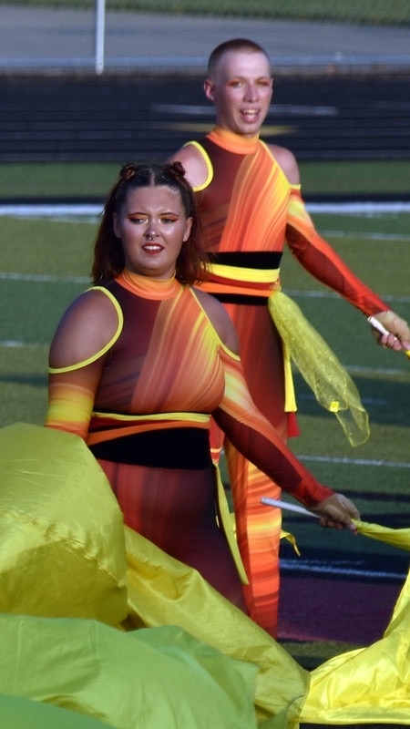 Two performers in bright, colorful costumes featuring orange and yellow patterns are dancing on a field. They are holding yellow flags and appear to be engaged in a performance, with a focus on movement and expression.