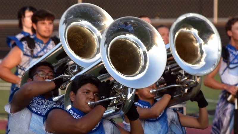 A group of musicians playing tubas during a performance, wearing blue and white uniforms. The focus is on three players in the foreground, with their instruments prominently displayed.