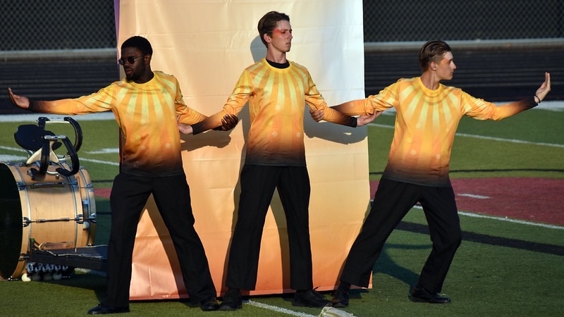 Three performers in brightly colored, gradient sunflower-patterned shirts stand in a line on a field, with one wearing sunglasses. They pose with arms extended, against a backdrop of a light-colored screen. A drum is visible in the foreground.