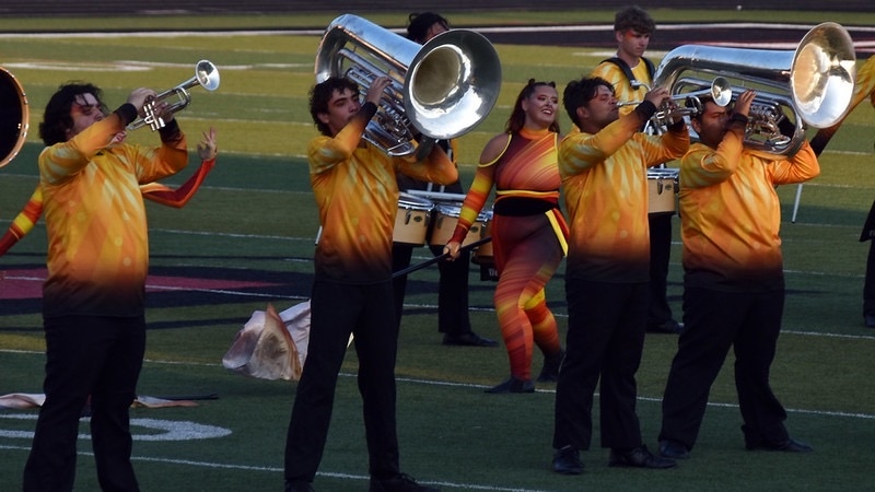 A drum corps performs on a field, with several musicians playing tubas. They wear vibrant, colorful uniforms that transition from yellow to orange. In the background, a member in a form-fitting costume adds to the ensemble's visual display.