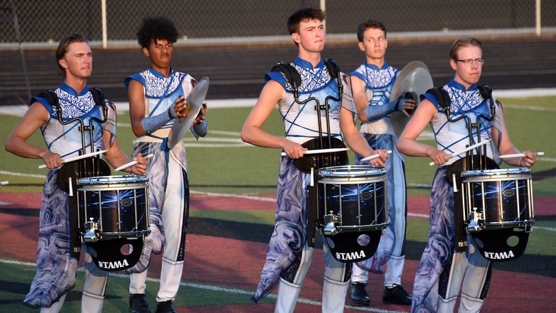 A group of five drummers in coordinated blue and white uniforms perform on a football field. They are playing drums and holding various percussion instruments, surrounded by turf and a track. The scene captures a moment of musical performance and teamwork.