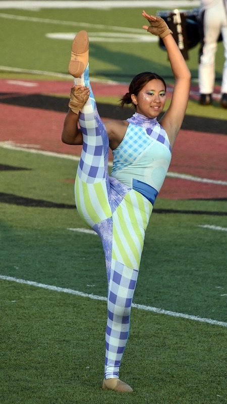 A dancer performs a high kick on a sports field, wearing a colorful, patterned costume. The background shows a blurred group of people in light-colored outfits, indicating a team or event. The setting is outdoors with green turf.