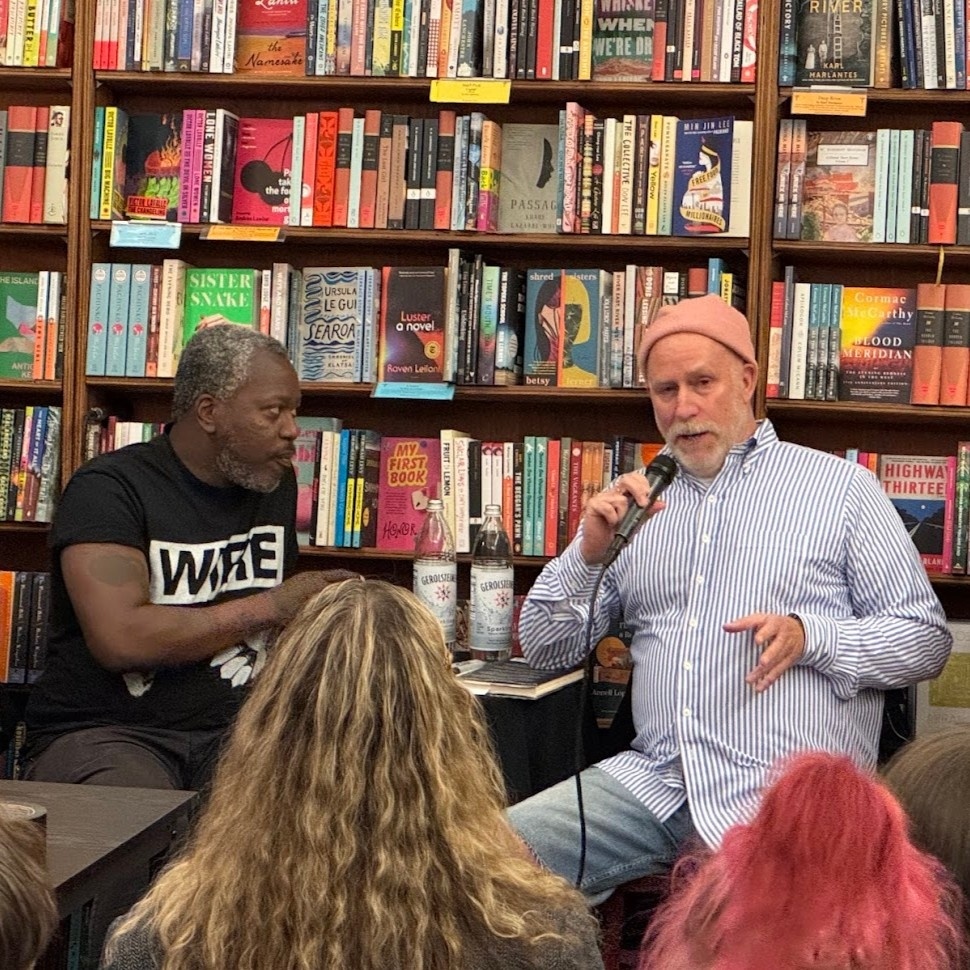 In City Lights. seated with shelves of books behind them, Roddy Bottum, in pink knit cap and blue and white buttoned shirt, speaks into a microphone while Brontez Purnell. in black Wire T-shirt, sits and looks at him on the left.
