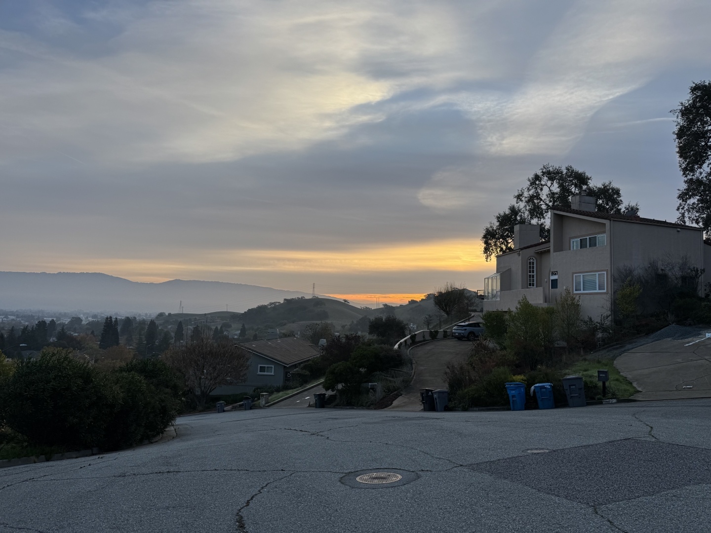 An early morning sunrise with some clouds, distant hills, nearby trees and houses and a road.