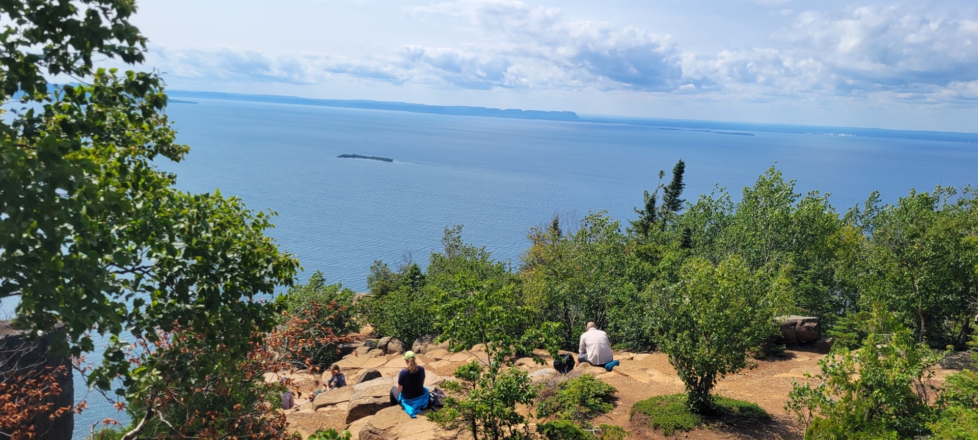 The lookout over Lake Superior with blue water and people sitting on red/brown rock, surrounded by trees.