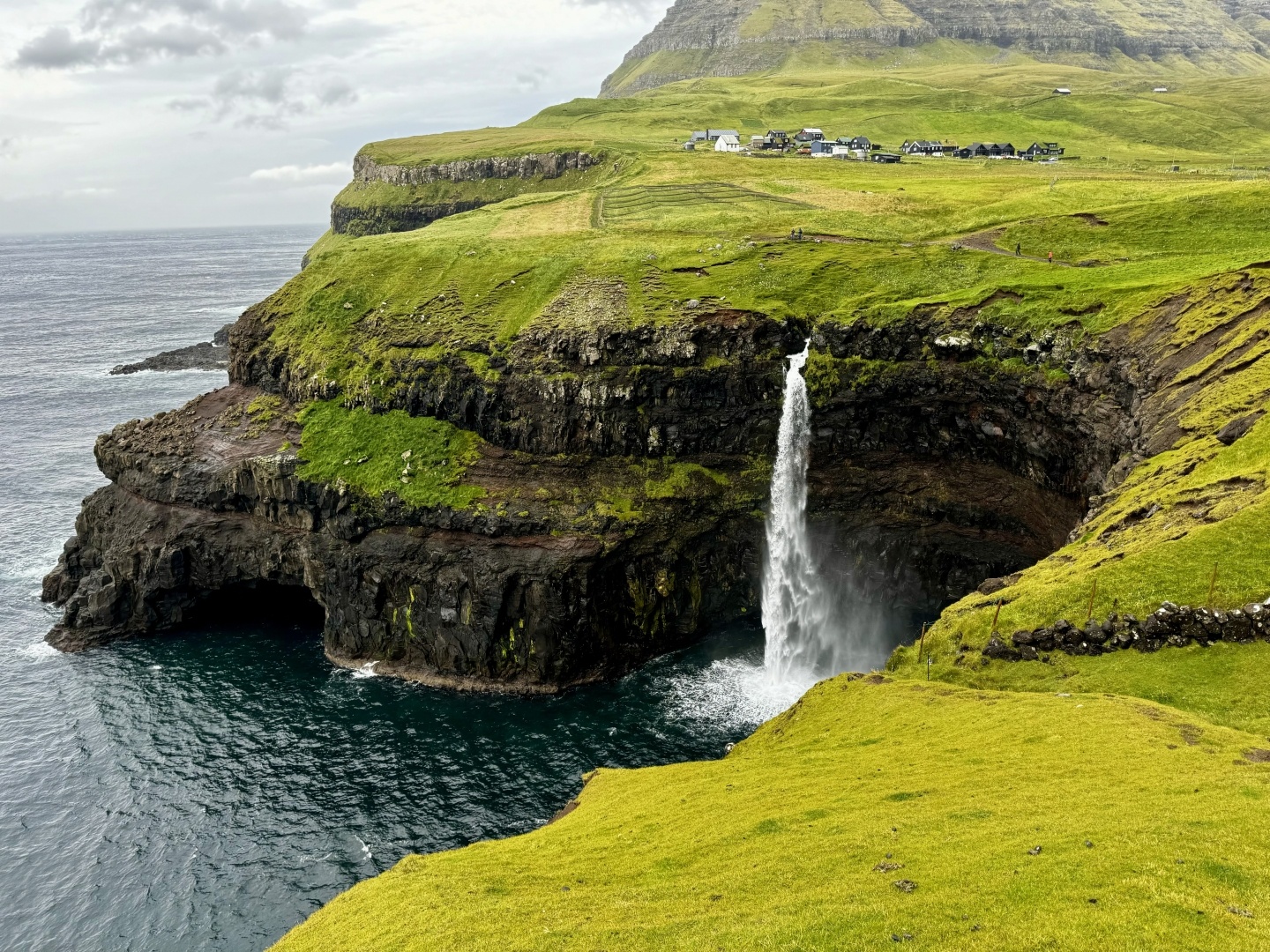 Picture of a waterfall falling to the sea, over a black rock cliff, surrounded by grass fields in a cloudy day