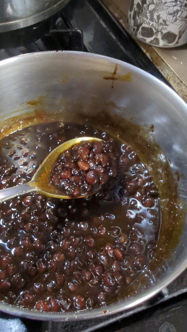 A metal pot of what appears to be baked beans, with a spoon holding up a scoop of them. They're in a thin, dark, sweet looking gravy.