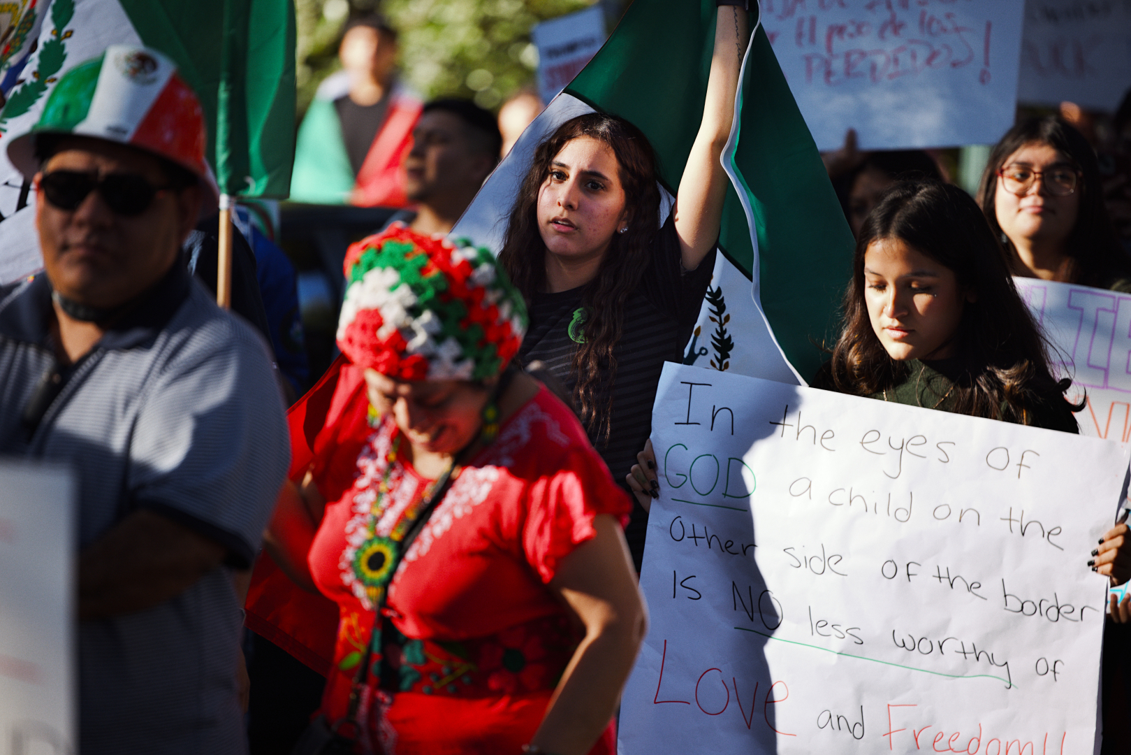 A group of people participate in a protest, holding signs and flags. A young woman in the center is looking at the camera while holding a Mexican flag in the sky. Another to her left holds a sign reading, "In the eyes of GOD, a child on the other side of the border is NO less worthy of LOVE and Freedom!" Another woman wears a colorful headdress and traditional clothing. The Mexican flag is visible in multiple instances among the crowd. The scene is lively and filled with sunlight.
