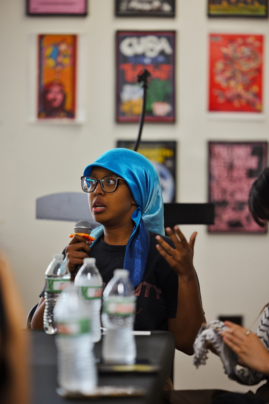 A person wearing glasses and a bright blue headscarf speaks into a microphone during a panel discussion. They are seated at a table with several water bottles in front. The background features a wall adorned with colorful posters. The person gestures with one hand while holding the microphone in the other, appearing engaged and expressive. Another person is partially visible to the right, focusing on something in their hands.