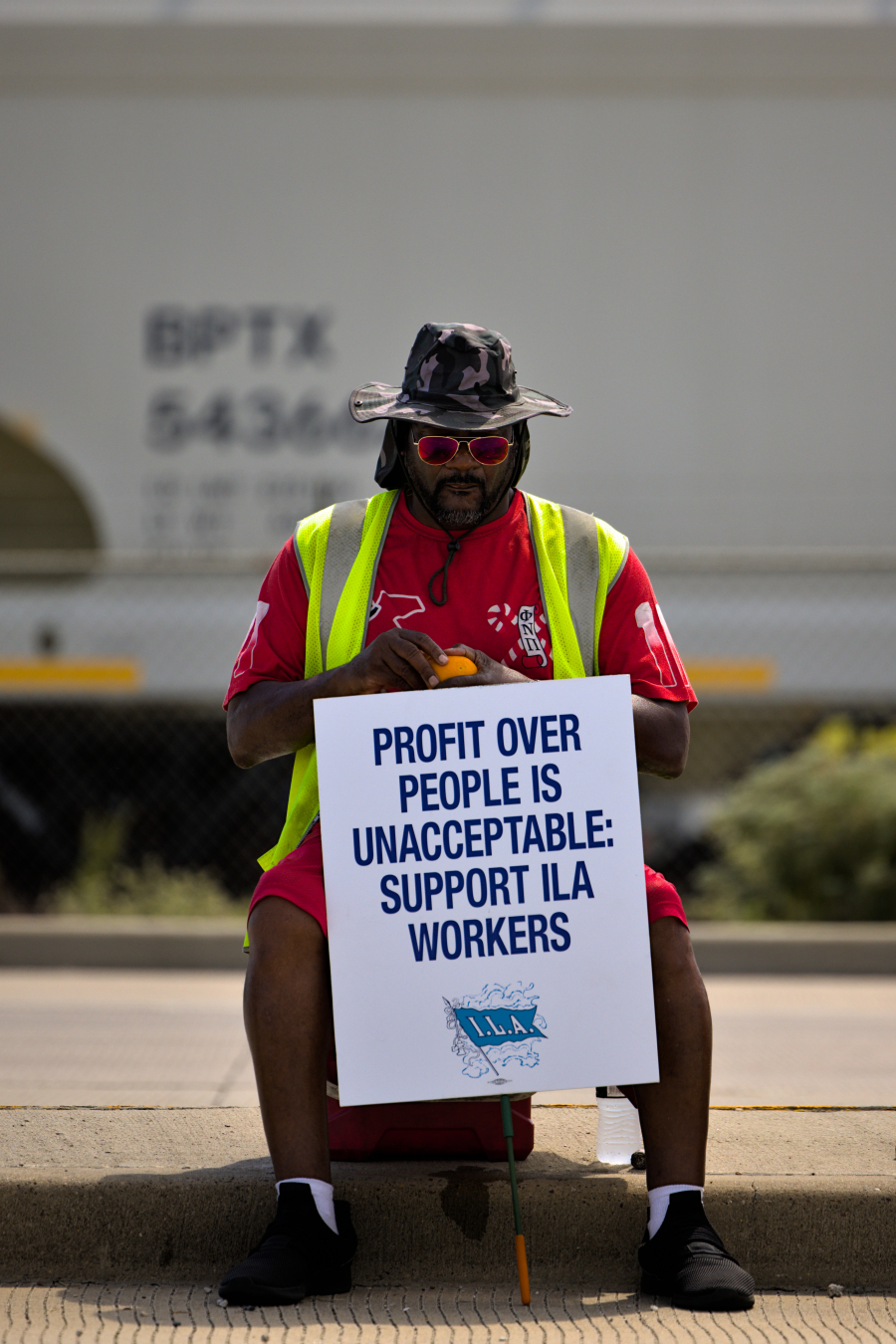 A person wearing a camouflage hat, sunglasses, and a neon safety vest sits on a cooler. They hold a sign reading "Profit over people is unacceptable: Support ILA workers" in bold blue letters. The individual wears a red shirt and black shoes, sitting on a sidewalk with a blurred industrial background. The scene suggests a protest or demonstration advocating for workers' rights.