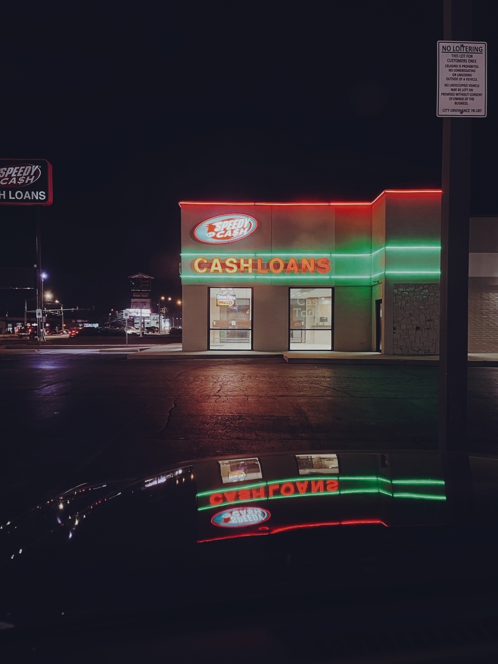 A photo of a title loan business at night. The sign is neon and is reflected of the hood of a vehicle.