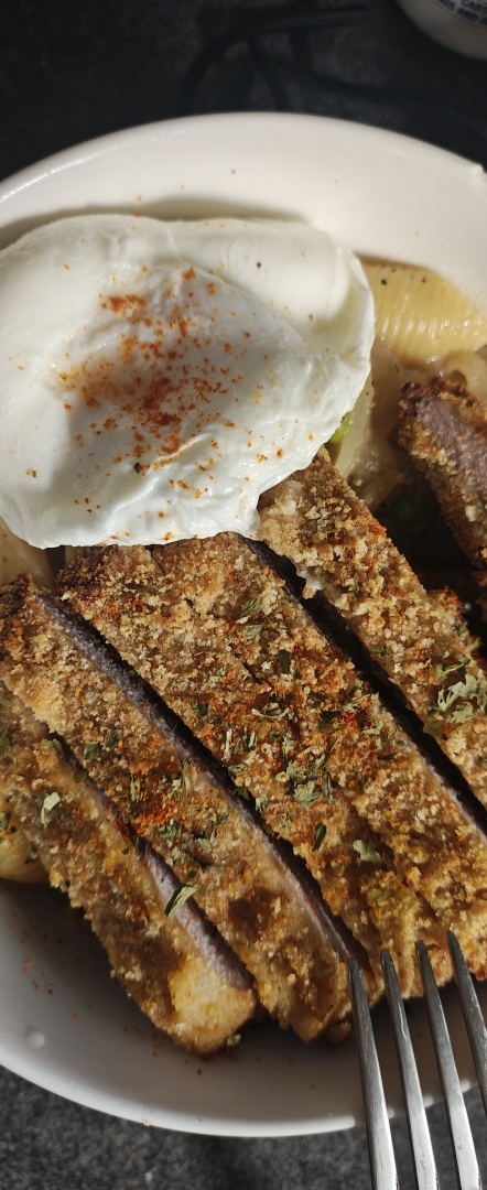 A top-down view of a meal featuring sliced, breaded meat cutlets seasoned with herbs and red spice, served alongside a poached egg sprinkled with paprika, over a bed of creamy shell pasta with a fork visible at the bottom.