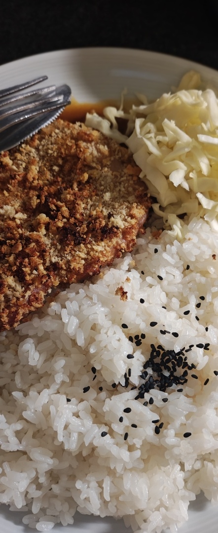 A close-up, overhead view of a meal served on a white plate. The dish features a golden-brown, breaded cutlet (likely katsu) on the left, accompanied by a pile of shredded cabbage on the top right. A large portion of white rice sprinkled with black sesame seeds fills the foreground, and a fork rests on the edge of the cutlet.